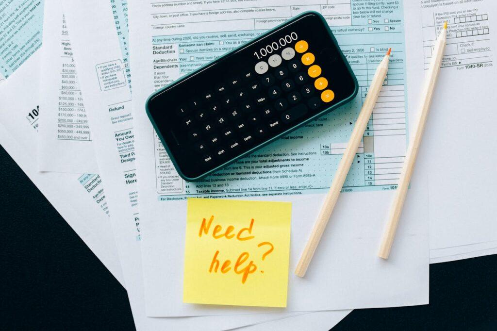 pexels-photo-6863260 Top view of financial papers with a calculator, pencils, and a note saying 'Need help?' indicating tax or accounting assistance.